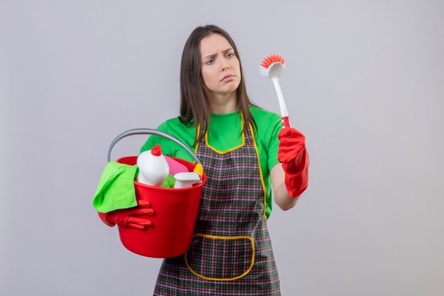 Triste limpieza joven con uniforme en guantes rojos sosteniendo herramientas de limpieza y mirando cepillo de limpieza en su mano sobre fondo blanco aislado