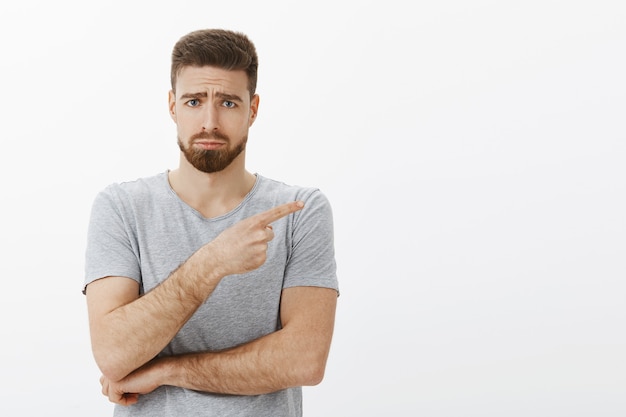 Foto gratuita triste y lindo modelo masculino barbudo encantador en camiseta gris frunciendo el ceño haciendo una cara sombría con el ceño fruncido, enfurruñado apuntando a la derecha expresando pesar y envidia de pie infeliz sobre la pared blanca