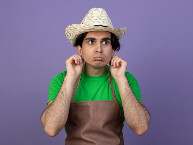 Triste mirando al lado joven jardinero en uniforme con sombrero de jardinería sosteniendo orejas aisladas en púrpura