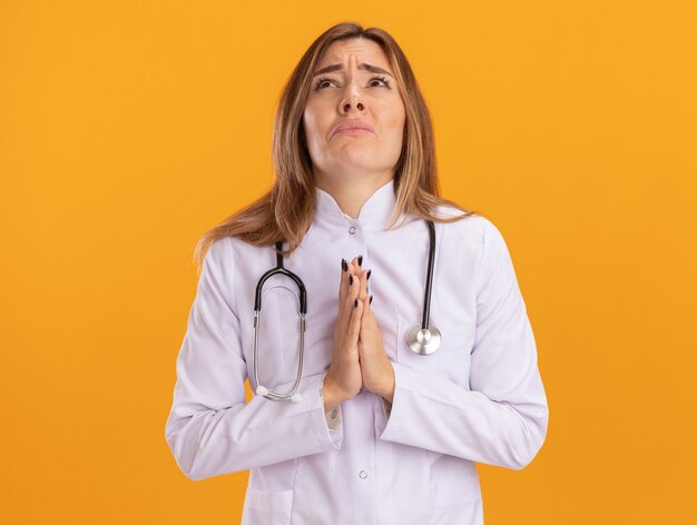 Triste mirando hacia arriba joven doctora vistiendo bata médica con estetoscopio mostrando gesto de oración aislado en la pared amarilla