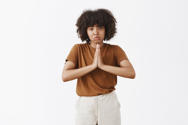 Triste molesta mujer adolescente de piel oscura y guapa con peinado afro frunciendo los labios con una sonrisa triste, frunciendo el ceño y tomados de la mano en oración mientras pide ayuda o favor sobre la pared gris