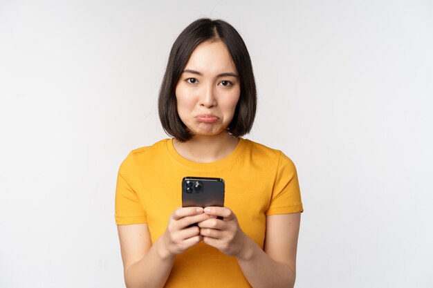 Triste mujer asiática sosteniendo un teléfono inteligente que parece molesto con arrepentimiento de pie en camiseta amarilla contra fondo blanco