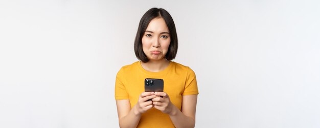 Triste mujer asiática sosteniendo un teléfono inteligente que parece molesto con arrepentimiento de pie en camiseta amarilla contra fondo blanco