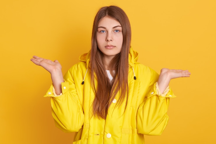Foto gratuito triste mujer caucásica extendiendo las manos a un lado, mostrando un gesto de impotencia, la señora con chaqueta, posando contra la pared amarilla, tiene una mirada seria, con expresión molesta.