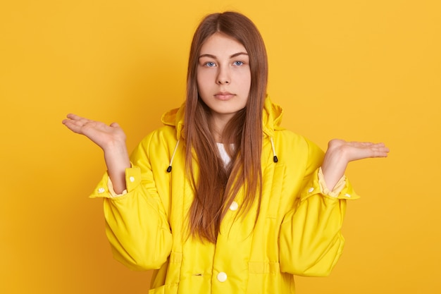 Triste mujer caucásica extendiendo las manos a un lado, mostrando un gesto de impotencia, la señora con chaqueta, posando contra la pared amarilla, tiene una mirada seria, con expresión molesta.