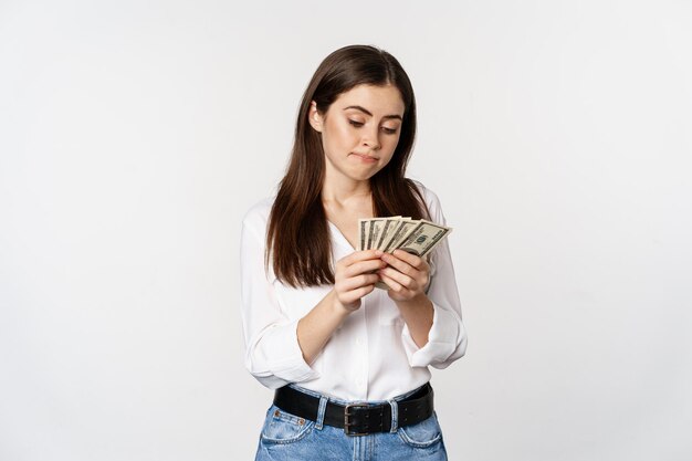 Triste mujer contando dinero, sin dinero en efectivo, de pie sombrío sobre fondo blanco.