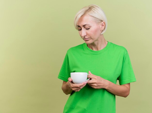 Triste mujer eslava rubia de mediana edad sosteniendo y mirando una taza de té aislado en la pared verde oliva con espacio de copia