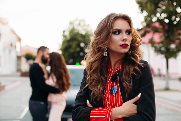 Triste mujer hermosa con los brazos cruzados en ropa de moda contra una pareja besándose.