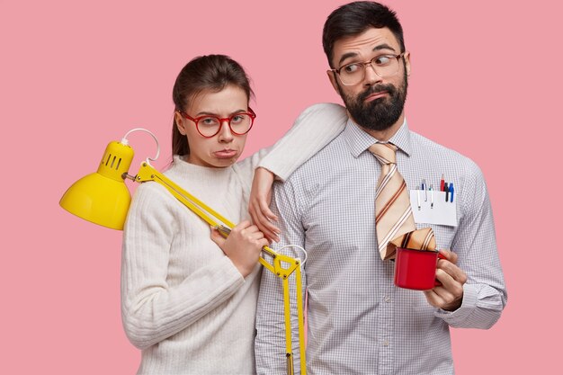 Triste mujer y hombre descontentos con gafas, hacen un trabajo común, sostienen la lámpara y la taza de café, expresan sentimientos negativos