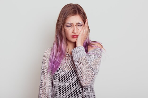 Triste mujer joven con cabello lila que sufre de un terrible dolor de muelas, tocándose la mejilla con la palma, mantiene los ojos cerrados, el ceño fruncido, aislado sobre fondo gris.