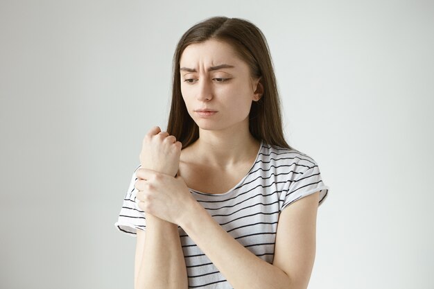 Triste mujer joven frustrada en la parte superior a rayas frunciendo el ceño, sosteniendo la mano en su muñeca adolorida, masajeando el área del dolor, con expresión facial dolorosa, sufriendo de dolor en las articulaciones, artritis o gota