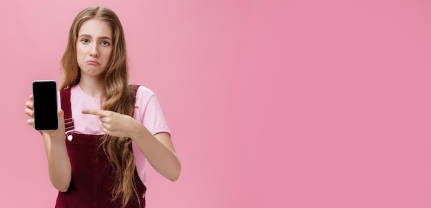 Foto gratuita triste mujer joven sombría con lindo peinado natural ondulado que muestra la pantalla del teléfono inteligente apuntando a gadge