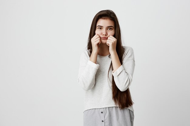 triste mujer morena guapa de ojos oscuros haciendo una mueca soplando sus mejillas y tirando de ellas fingiendo divertirse. Hermosa mujer vestida con ropa casual se ofende