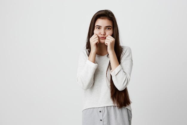 triste mujer morena guapa de ojos oscuros haciendo una mueca soplando sus mejillas y tirando de ellas fingiendo divertirse. Hermosa mujer vestida con ropa casual se ofende