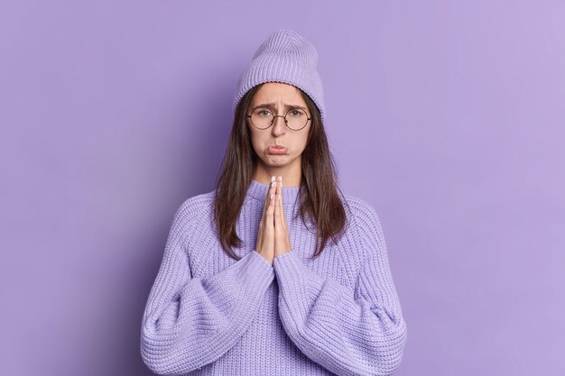 Triste mujer morena mira con expresión suplicante mantiene las palmas juntas pide disculpas bolsos labios usa gafas redondas vestidas con suéter de punto y sombrero.