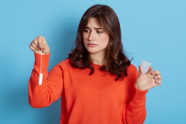 Triste mujer de pelo oscuro vistiendo un suéter naranja sosteniendo en las manos productos higiénicos de las mujeres