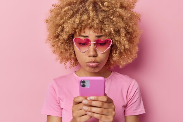 Triste mujer de pelo rizado frustrada mira infelizmente la pantalla del teléfono inteligente se siente arrepentida usa gafas de sol en forma de corazón camiseta casual aislada sobre fondo rosa cheques cuenta bancaria se siente sola