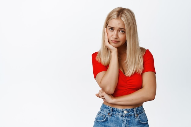 Triste mujer rubia cabeza inclinada en la cara con aspecto molesto y malhumorado de pie en la parte superior recortada y jeans contra fondo blanco Copiar espacio