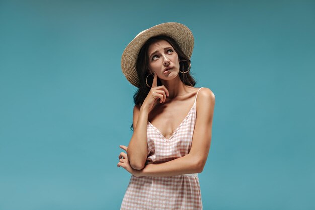 Triste mujer de verano con el pelo largo en un elegante sombrero de ala ancha y un vestido rosa y blanco a cuadros mirando hacia otro lado en un contexto aislado