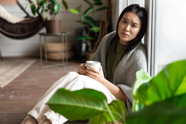 Triste niña asiática enferma sentada con taza de café cerca de la ventana en el piso con aspecto molesto e infeliz