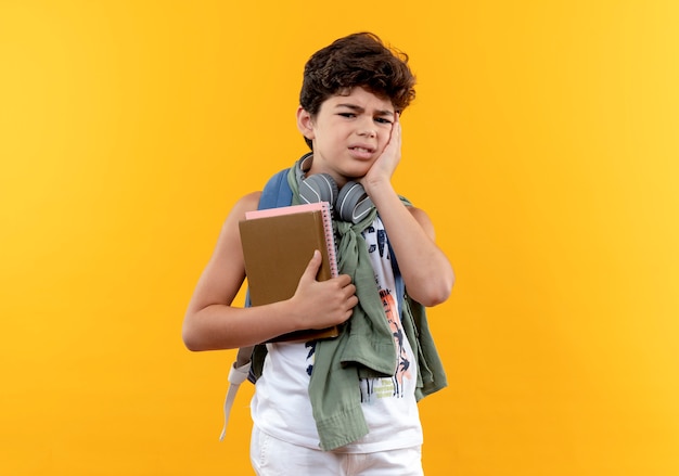 Triste niño de escuela con mochila y auriculares sosteniendo libros y poniendo la mano en la mejilla aislada en amarillo