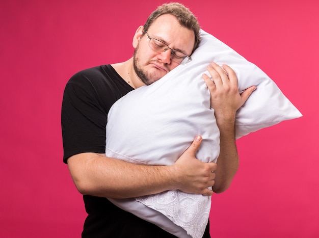 Triste con los ojos cerrados, varón enfermo de mediana edad abrazó la almohada aislada en la pared rosada