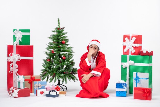 Triste pensativo joven vestido como Papá Noel con regalos y árbol de Navidad decorado sentado en el suelo sobre fondo blanco.