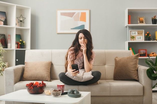 Foto gratuita triste poner el dedo en los labios niña sosteniendo el teléfono sentado en el sofá detrás de la mesa de café en la sala de estar