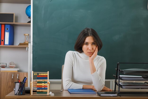 triste poner la mano en la mejilla joven maestra sentada en el escritorio con herramientas escolares en el aula