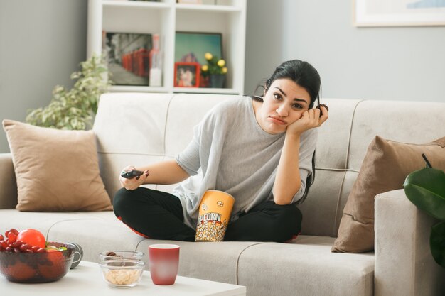 Triste poner la mano en la mejilla niña sosteniendo el control remoto del televisor con galleta sentada en el sofá detrás de la mesa de café en la sala de estar