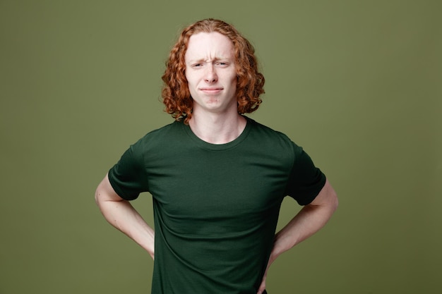 Triste poner las manos en las caderas joven guapo con camiseta verde aislado sobre fondo verde