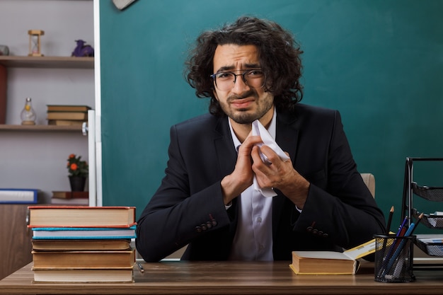 Triste profesor con gafas triturando papel sentado a la mesa con herramientas escolares en el aula