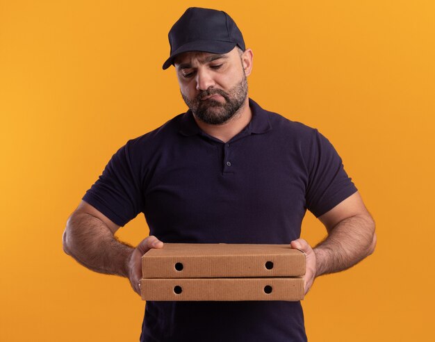 Triste repartidor de mediana edad en uniforme y gorra sosteniendo y mirando cajas de pizza aisladas en la pared amarilla
