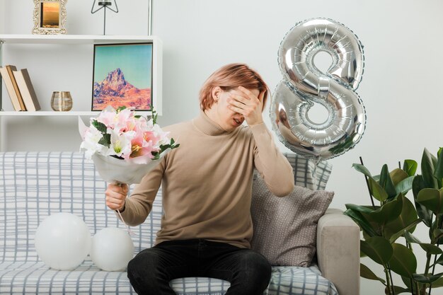 Triste rostro cubierto con la mano chico guapo en el día de la mujer feliz con ramo sentado en el sofá en la sala de estar