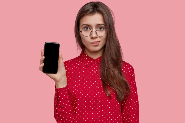 Triste señorita frunce los labios, sostiene un teléfono celular moderno con pantalla de maqueta, usa gafas transparentes, vestida con una camisa de lunares