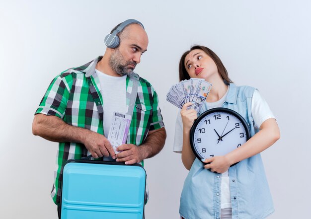 Triste viajero adulto pareja hombre usando audífonos sosteniendo boletos de viaje y maleta mujer sosteniendo dinero y reloj mirando el uno al otro