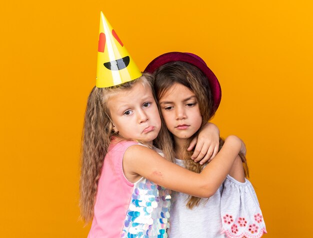 Tristes niñas bonitas con sombreros de fiesta abrazándose unos a otros aislados en la pared naranja con espacio de copia