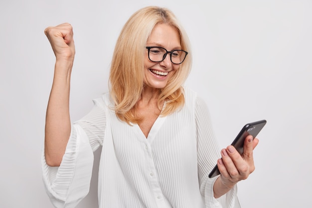 Triunfante mujer de mediana edad rubia positiva aprieta el puño de alegría celebra el éxito lee noticias increíbles a través del teléfono inteligente recibió comentarios positivos usa gafas y blusa aislada en la pared blanca