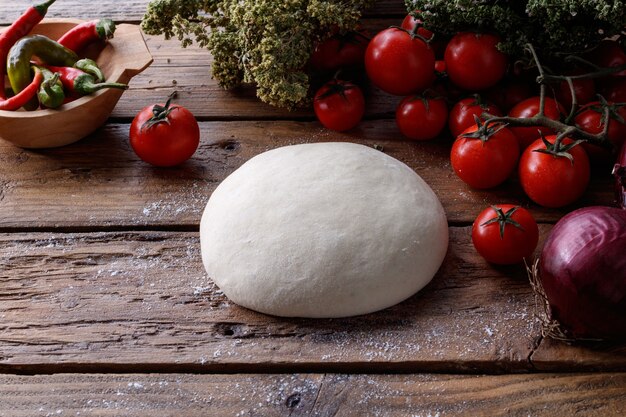 Trozo de masa sobre una mesa de madera rodeada de tomates, pimiento y cebolla