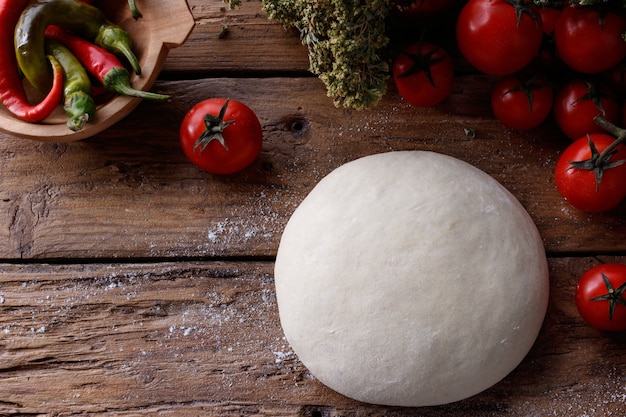 Trozo de masa sobre una mesa de madera rodeada de tomates y pimiento