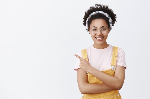 Tu camino hasta allí. Retrato de afroamericano confiado y feliz de aspecto amistoso con gafas, diadema de moda y monos amarillos, apuntando a la esquina superior izquierda y sonriendo alegremente sobre la pared gris