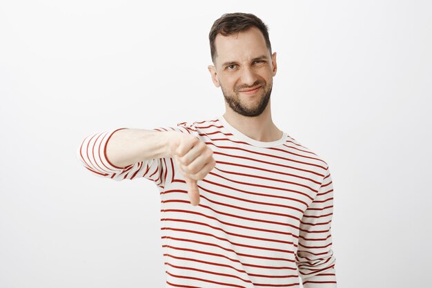 Tu idea apesta. Retrato de hombre guapo disgustado no impresionado con barba, haciendo muecas de disgusto y mostrando los pulgares hacia abajo, frunciendo el ceño