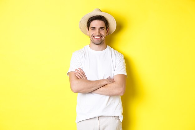 Turismo. Hombre joven guapo que parece feliz, con sombrero de paja para viajar, cruzando las manos sobre el pecho y sonriendo, de pie sobre un fondo amarillo.