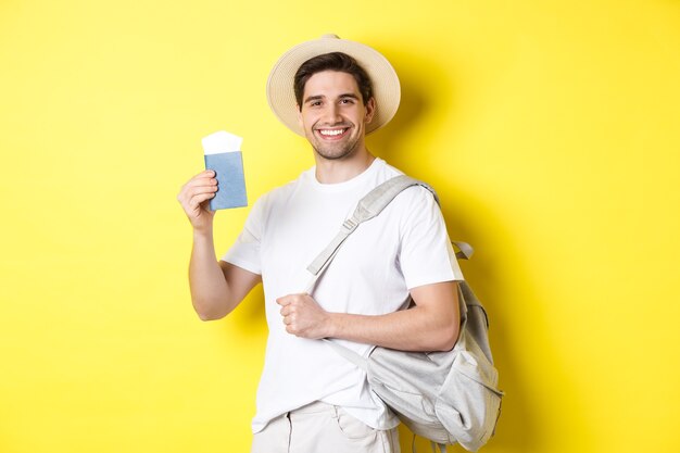 Turismo y vacaciones. Chico joven sonriente que va de viaje, sosteniendo la mochila y mostrando el pasaporte con boletos, de pie sobre un fondo amarillo.