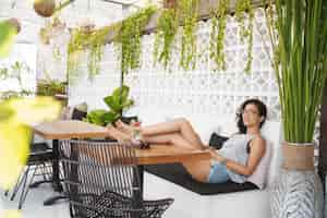 Foto gratuita turismo de vacaciones y concepto de ocio feliz mujer morena bronceada atractiva y relajada con gafas tumbadas en la zona de salón del hotel terraza del café poner los pies en la mesa de café sintiéndose sonriente en el paraíso