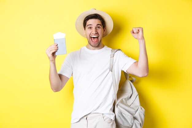 Turismo y vacaciones. El hombre se siente feliz por el viaje de verano, sosteniendo el pasaporte con los boletos de avión y la mochila, levantando las manos en gesto de celebración, fondo amarillo