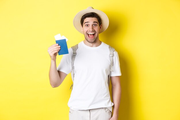 Turismo y vacaciones. Turista hombre feliz mostrando su pasaporte con boletos, yendo de viaje, de pie sobre fondo amarillo con mochila