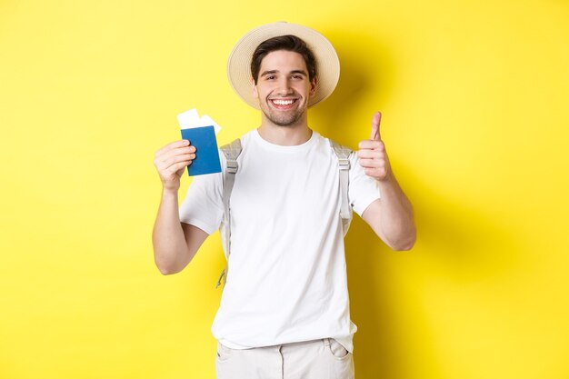 Turismo y vacaciones. Turista masculino satisfecho mostrando pasaporte con boletos y pulgar hacia arriba, recomendando la compañía de viajes, de pie sobre fondo amarillo