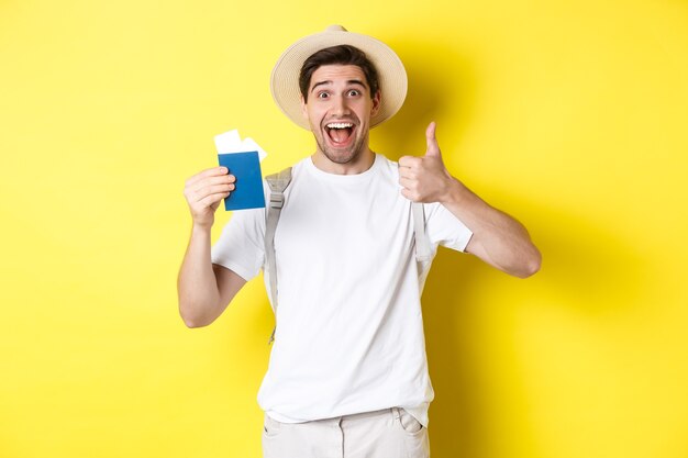 Turismo y vacaciones. Turista masculino satisfecho que muestra el pasaporte con los billetes y el pulgar hacia arriba, recomendando la compañía de viajes, de pie sobre un fondo amarillo.