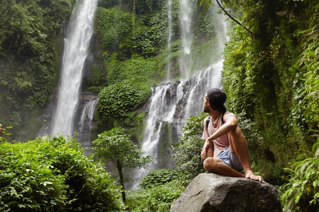 Turismo, viajes y aventura. Elegante joven inconformista sentado en piedra con los pies descalzos y volviendo la cabeza hacia atrás para ver una cascada increíble